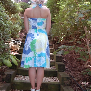 May include: A strapless knee-length dress featuring a white base with a large-scale floral print in shades of blue, green, and yellow. The dress includes a matching fabric hair tie and is paired with white block-heeled sandals with ankle straps.