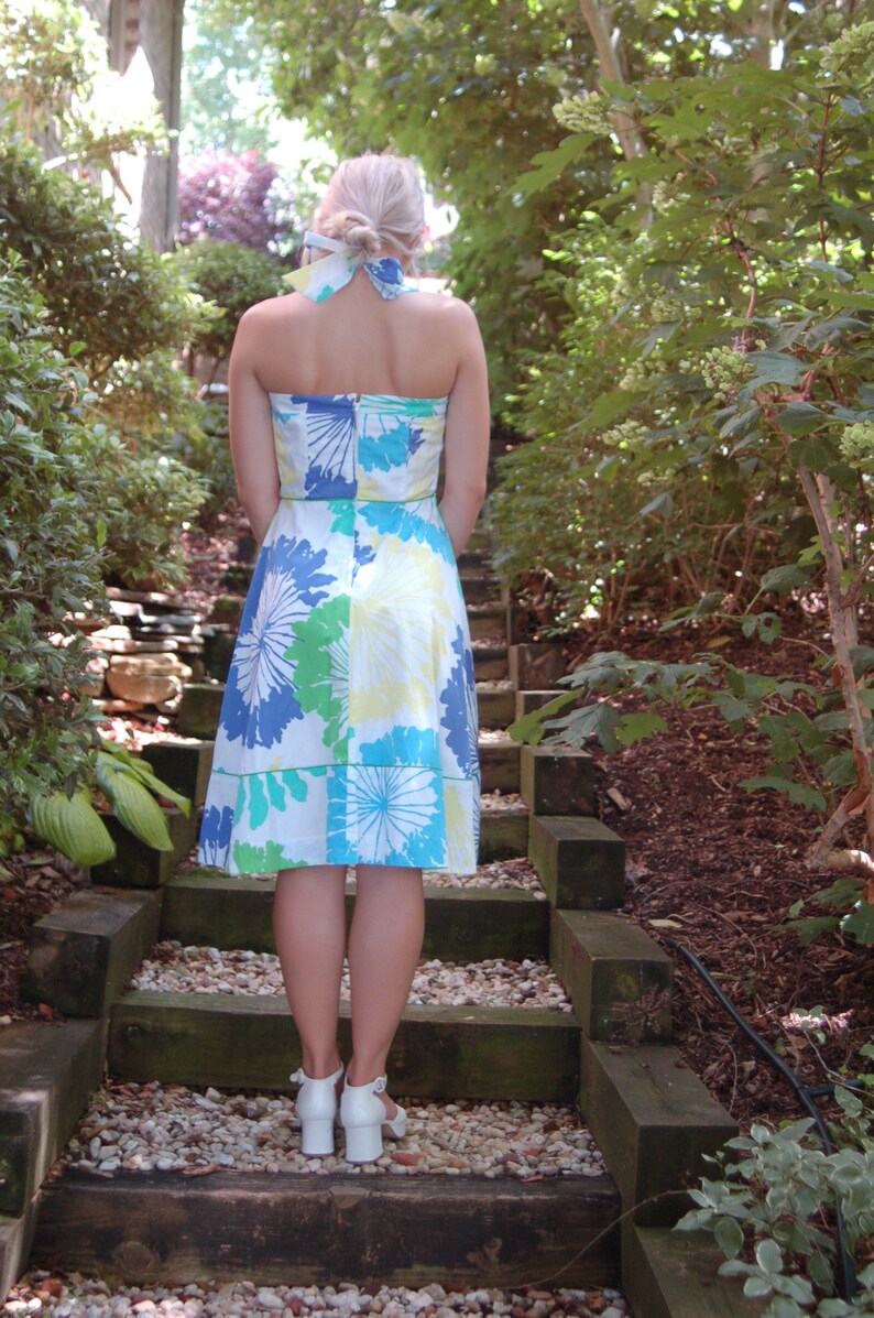 May include: A strapless knee-length dress featuring a white base with a large-scale floral print in shades of blue, green, and yellow. The dress includes a matching fabric hair tie and is paired with white block-heeled sandals with ankle straps.