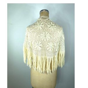 May include: Cream-colored lace shawl with a floral pattern and fringe. The shawl is draped over a mannequin, showcasing the intricate details of the lace design. The fringe hangs along the bottom edge.