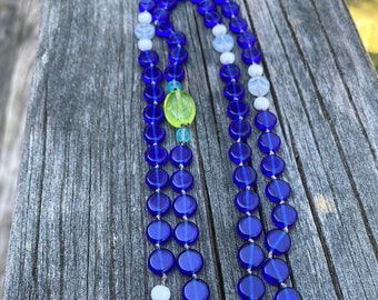 May include: A blue and white glass bead necklace with a green bead accent. The necklace is made of round beads and is strung on a cord.