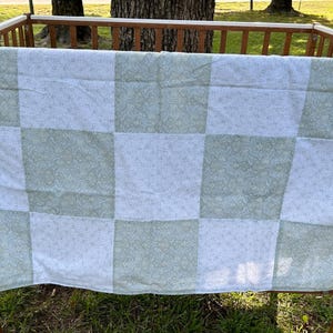 May include: A white and green checkered baby blanket with a floral pattern. The blanket is hanging on a wooden crib.