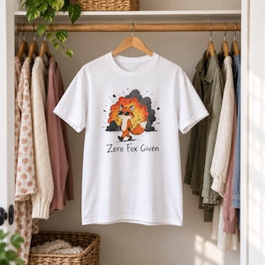 May include: A white short-sleeved t-shirt featuring a graphic illustration of an orange fox wearing black sunglasses walking away from a large orange and black explosion. The text "Zero Fox Given" is printed in black below the fox illustration.