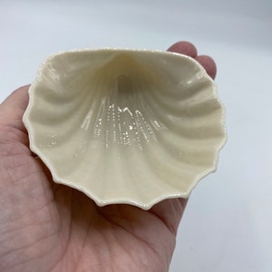 Puede incluir: Un pequeño plato de cerámica blanco con forma de concha. El plato tiene un acabado liso y brillante y un borde ligeramente festoneado.