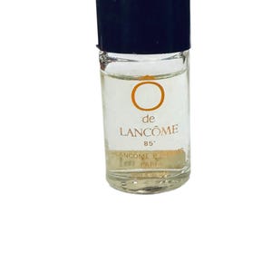 Könnte beinhalten: Eine kleine, klare Glasparfümflasche mit schwarzem Verschluss. Die Flasche hat ein Etikett mit den Aufschriften "de LANCÔME" und "85". Die Flasche ist mit einer klaren Flüssigkeit gefüllt.