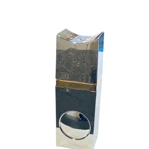 Puede incluir: Un objeto rectangular plateado con una parte superior curvada y un recorte circular. El objeto tiene una banda dorada y un acabado espejado, que refleja el entorno. El diseño es moderno y geométrico.