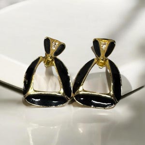 May include: A pair of gold-tone earrings with black enamel and clear rhinestones. The earrings are shaped like a triangle with a small circle in the center.
