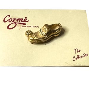 May include: Gold-toned shoe-shaped pin with a golf ball on the toe. The pin is on a card that reads "Cozme International" and "The Collection".