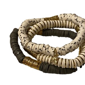 Könnte beinhalten: Ein Set aus drei Armbändern mit weißen, schwarzen und goldenen Perlen. Die weißen Perlen haben ein schwarz-weißes Muster. Die goldenen Perlen sind klein und rund. Die Armbänder sind übereinander gestapelt.