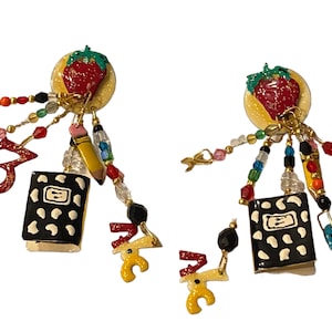 May include: A pair of dangle earrings with a school theme. Each earring features a red strawberry, a black and white book with white polka dots, a yellow pencil, a red number 3, and a variety of colourful beads.
