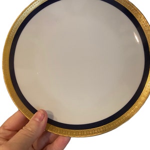 以下が含まれることがあります： 金と青の縁取りが施された白い磁器の皿。縁取りには、連なった円が繰り返される模様が施されています。