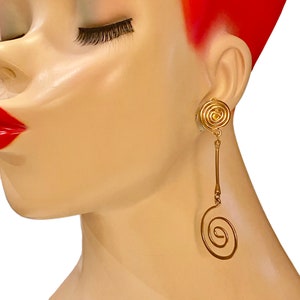 Puede incluir: Pendientes de aro dorados con diseño de espiral. Los pendientes son largos y tienen una pequeña espiral en la parte superior y una espiral más grande en la parte inferior.