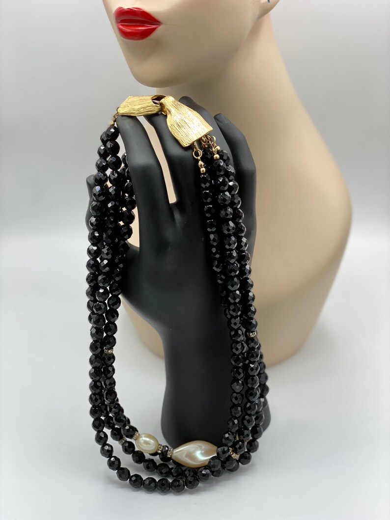 Pu&ograve; includere: Una collana multifilo realizzata con perline nere, con una chiusura dorata e un accento di perla bianca.