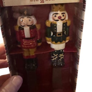 May include: A boxed set of two Nutcracker spreaders from Williams Sonoma. The spreaders are shaped like toy soldiers, one in red and gold, the other in green and gold. The box has the words "Nutcracker Spreaders" and "Set of two".