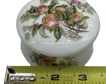 Könnte beinhalten: Weiße Keramik-Schmuckdose mit Deckel. Der Deckel ist mit einem Blumendesign verziert, das rosa und grüne Blumen und Blätter zeigt. Die Dose hat einen Durchmesser von etwa 5 cm.