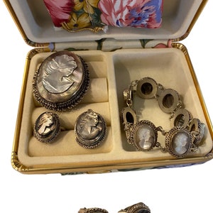 Könnte beinhalten: Ein Set Kameen-Schmuck in einer Schachtel mit Blumenmuster. Das Set enthält eine Brosche, ein Armband und ein Paar Ohrringe. Der Schmuck ist aus Silber und zeigt ein Kameen-Design mit dem Profil einer Frau.