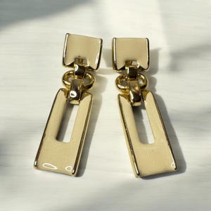 Puede incluir: Un par de pendientes colgantes de color dorado y crema con un diseño rectangular. Los pendientes presentan un eslabón de cadena dorado y un colgante rectangular de color crema.