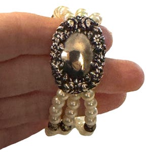 May include: An antique bracelet featuring faux pearls and an oval, silver-toned centerpiece. The centerpiece is surrounded by a decorative border. The bracelet has three strands of small, off-white pearls.
