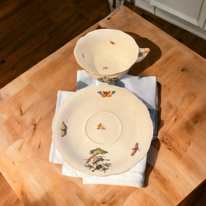 Puede incluir: Una taza de té y un platillo de porcelana blanca con un delicado diseño floral y de mariposas. La taza y el platillo están sobre una servilleta blanca en una mesa de madera.