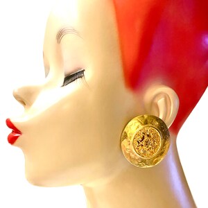 May include: A pair of large, gold-toned earrings with a circular, openwork design. The earrings are shown on a mannequin's ear.