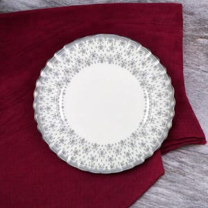 Op de afbeelding: Een wit keramiek bord met een gegolfde rand en een grijs bloemenpatroon. Het bord ligt op een bordeauxrode doek.