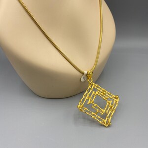 Puede incluir: Un collar de cadena de oro con un gran colgante cuadrado hecho de barras de oro entrelazadas. El colgante tiene un diseño geométrico.
