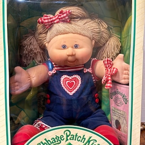 May include: A Cabbage Patch Kids doll with blonde yarn hair in pigtails and a red and white gingham bow. The doll wears a navy blue corduroy overall dress with a red heart graphic, a red and white striped shirt, and red shoes in a box.