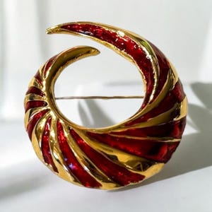 May include: A gold-toned brooch with a red enamel swirl design. The brooch is shaped like a crescent moon and has a pin on the back.