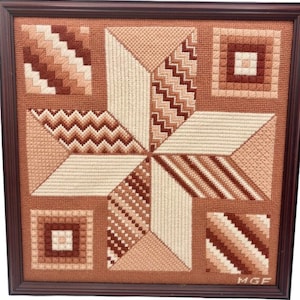 May include: A framed needlepoint piece with a starburst design in shades of brown and beige. The piece is signed "MGF" in the bottom right corner.