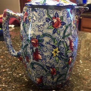 May include: A blue and white ceramic teapot with a floral pattern. The teapot has a lid with a knob on top. The floral pattern includes red, yellow, and green flowers.