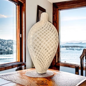 May include: White glass vase with a woven design. The vase is sitting on a wooden table in front of a window with a view of a body of water and mountains.