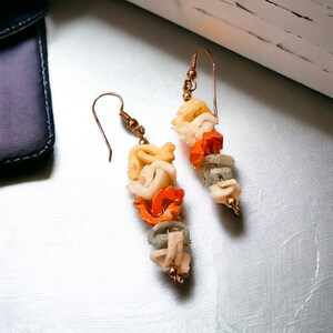 Puede incluir: Un par de pendientes de oro con formas de pasta de cerámica blanca, naranja y gris. Los pendientes son colgantes y tienen un cierre de gancho de oro simple.