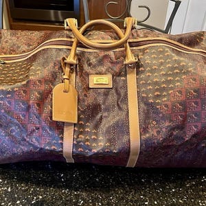 May include: A large brown duffel bag featuring a repeating geometric and floral pattern in shades of dark red, orange, and black. The bag includes tan leather handles, matching trim, a gold-toned zipper, and a rectangular luggage tag attached.
