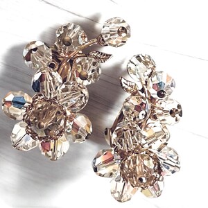 May include: A pair of vintage clip-on earrings featuring clusters of faceted, clear crystal beads. The beads are arranged in a floral design with gold-toned metal stems and leaves. The earrings have a classic, elegant style.