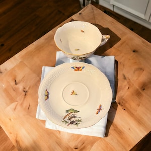 Puede incluir: Una taza de té y un platillo de porcelana blanca con un diseño floral y de mariposas. La taza tiene un borde festoneado y un asa. El platillo tiene un borde elevado y un patrón delicado.