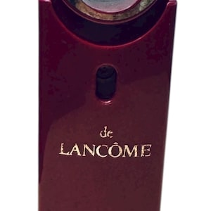 Puede incluir: Frasco de perfume burdeos con un anillo plateado en la parte superior. El frasco tiene las palabras "de LANCÔME" y "LANCÔME PARIS" impresas. El diseño es elegante y moderno.