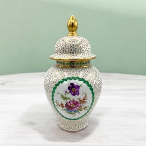Może przedstawiać: Biały porcelanowy dzbanek z pozłacaną pokrywką i kwiatowym wzorem. Dzbanek ma białe tło z złotym i zielonym wzorem. Kwiatowy wzór znajduje się w środku dzbanka i przedstawia fioletowe, różowe i żółte kwiaty.