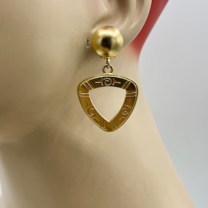 May include: Gold tone clip-on earrings with a triangular pendant featuring a geometric design. The pendant has a circular opening in the center.