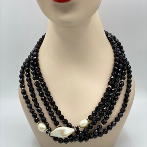 Pu&ograve; includere: Una collana multifilo di perle nere con un ciondolo a conchiglia bianca di grandi dimensioni. La collana &egrave; realizzata con perle nere sfaccettate ed &egrave; stratificata per un look drammatico.