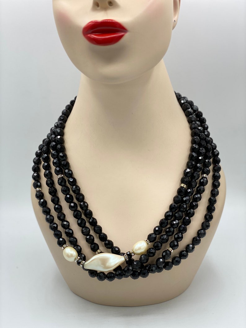 Pu&ograve; includere: Una collana multifilo di perle nere con un ciondolo a conchiglia bianca di grandi dimensioni. La collana &egrave; realizzata con perle nere sfaccettate ed &egrave; stratificata per un look drammatico.