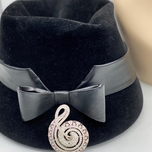 May include: A black velvet hat with a black leather band and bow. A silver treble clef brooch with rhinestones is attached to the bow.