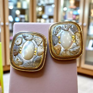 May include: A pair of square-shaped earrings with a gold-colored border. Each earring features a central oval-shaped, pearlescent stone surrounded by small, irregularly shaped stones in shades of gray, beige, and brown.