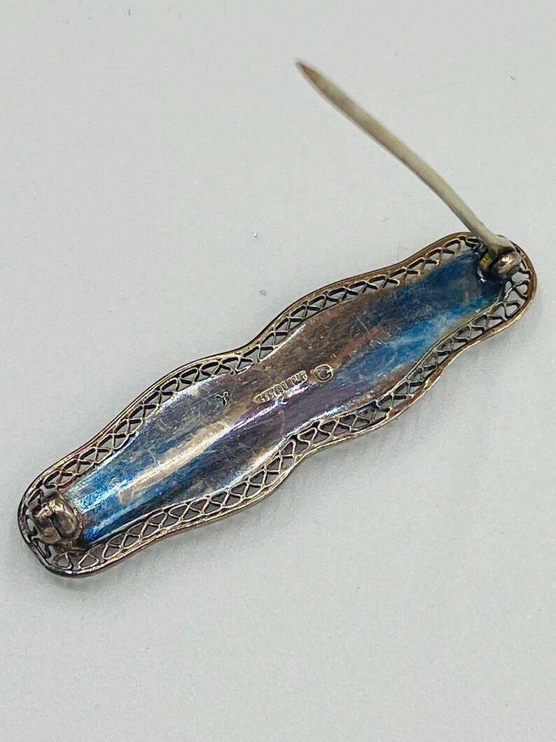 May include: A silver filigree brooch with a pin on the back. The brooch is marked "STERLING" on the back.