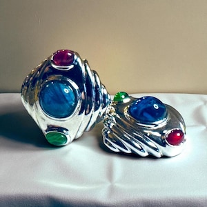 May include: Two silver-colored, ornate rings with colorful gemstone accents. One ring features a large blue stone, a red stone, and a green stone. The other ring has a blue stone and a red stone. The rings are on a white surface.