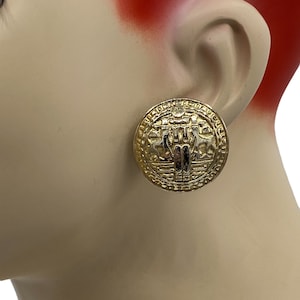 Puede incluir: Pendientes de clip en tono dorado con un diseño circular que presenta un patrón detallado en relieve. El centro de los pendientes presenta un diseño estilizado en relieve.