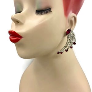 Puede incluir: Un pendiente plateado con un diseño en cascada, con pedrería transparente y roja. El pendiente se lleva en una cabeza de maniquí con pintalabios rojo y peluca roja.