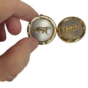 以下が含まれることがあります： 銀色のセンターを持つ金色のカフスボタンのペア。各カフスボタンには、スタイリッシュなパンサーの画像が描かれています。
