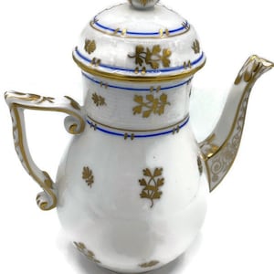 May include: A white porcelain creamer with a gold and blue floral design. The creamer has a lid and a handle.