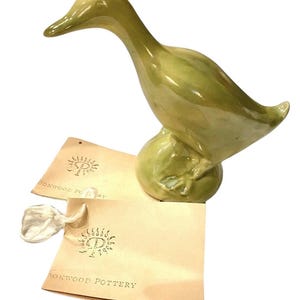 May include: A ceramic duck figurine in shades of green, perched on a rounded base. The duck has a long neck and a detailed beak. Two tags with the text "DORKWOOD POTTERY" are included.