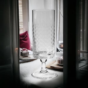 May include: A clear glass vase with a textured, patterned design. The vase has a tall, cylindrical body and a wide, flared base.