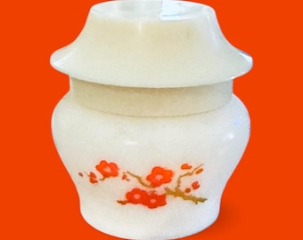 Puede incluir: Un pequeño jarrón blanquecino con tapa, con un diseño floral de flores rojas sobre una rama. El jarrón tiene un cuerpo redondeado y una parte superior ensanchada, sobre un fondo naranja. El jarrón es probablemente de piedra.
